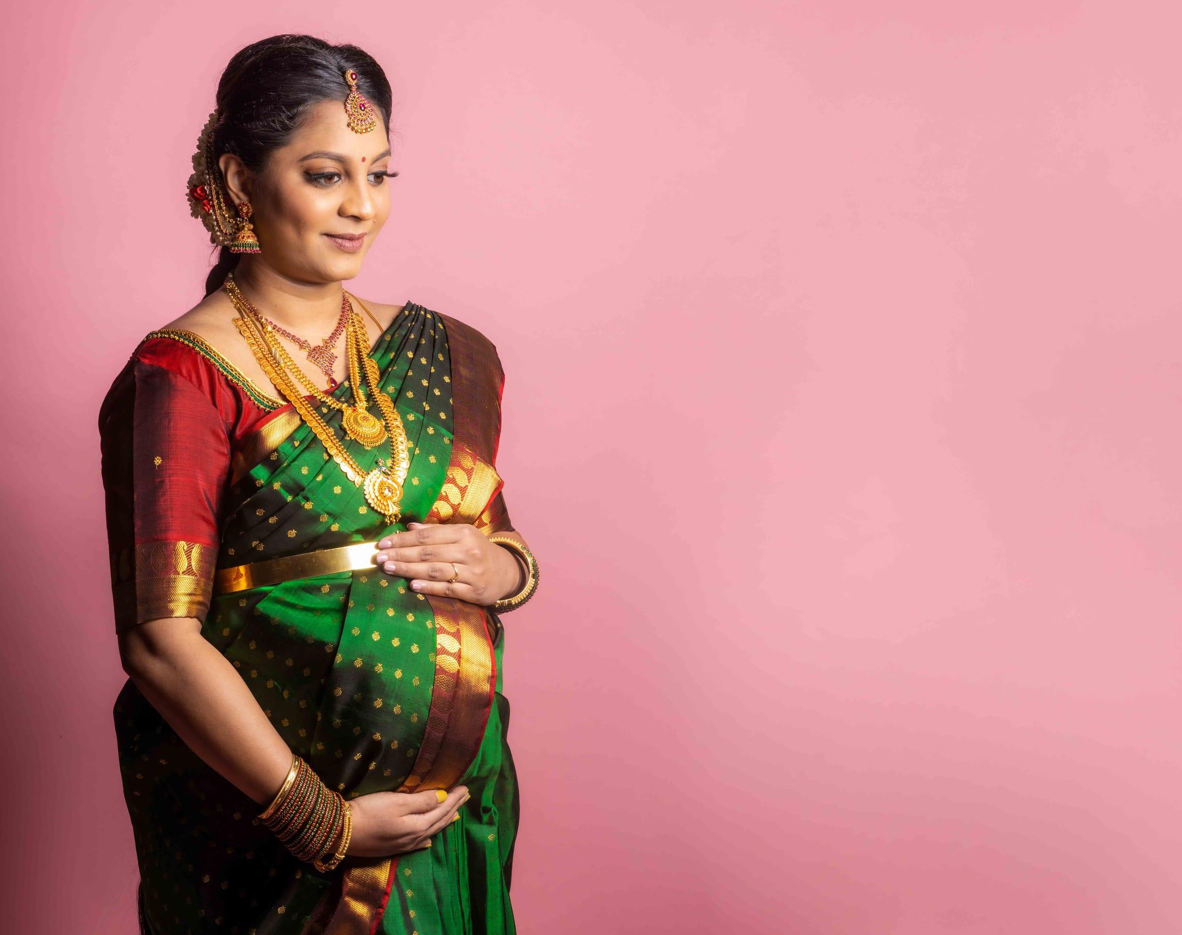 Expectant mother in a green silk saree on a pink backdrop, studio maternity portrait