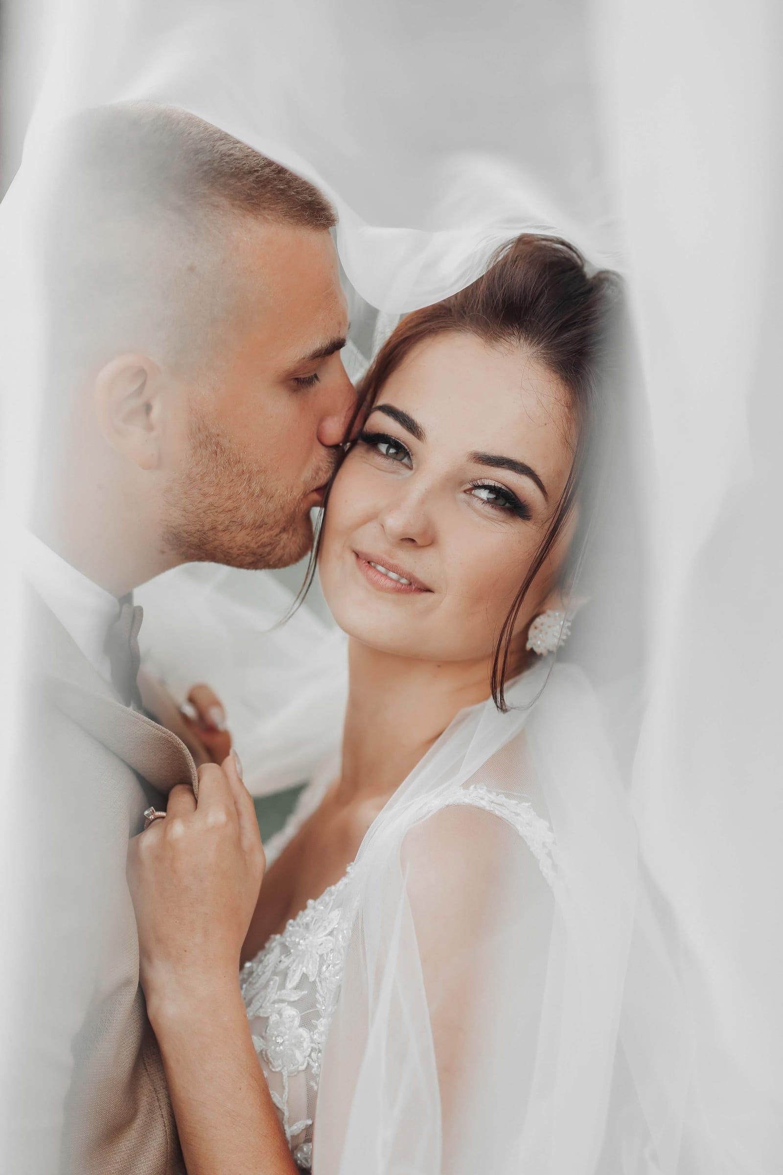 Groom kisses bride's cheek beneath a flowing veil, timeless wedding portrait